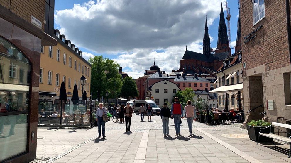 Personer går eller sitter vid uteserveringar på Gamla torget i Uppsala.