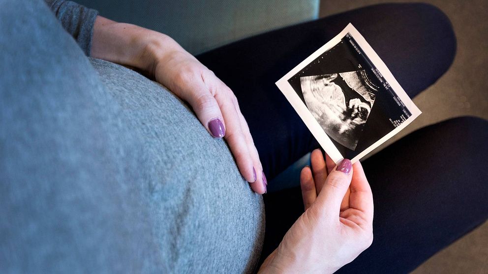 närbild ner mot en kvinnas gravidmage, ena handen på magen, andra håller en ultraljudsbild