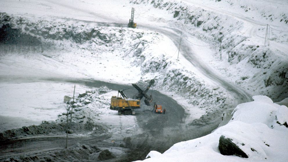 en gruvmaskin, en bil och en människa syns nere i en jättelik grusgrop med en väg ner, tunt snötäcke
