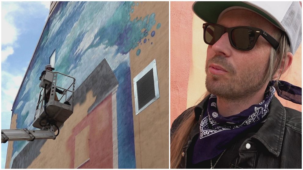 Till vänster en skylift med en konstnär som håller på att göra en muralmålning på en husvägg. Till höger en man med svarta glasögon och vit keps.