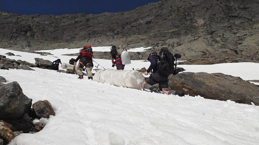 flera personer med ryggsäckar drar två stora plastade rullar uppför en brant sluttning med snö och klippor