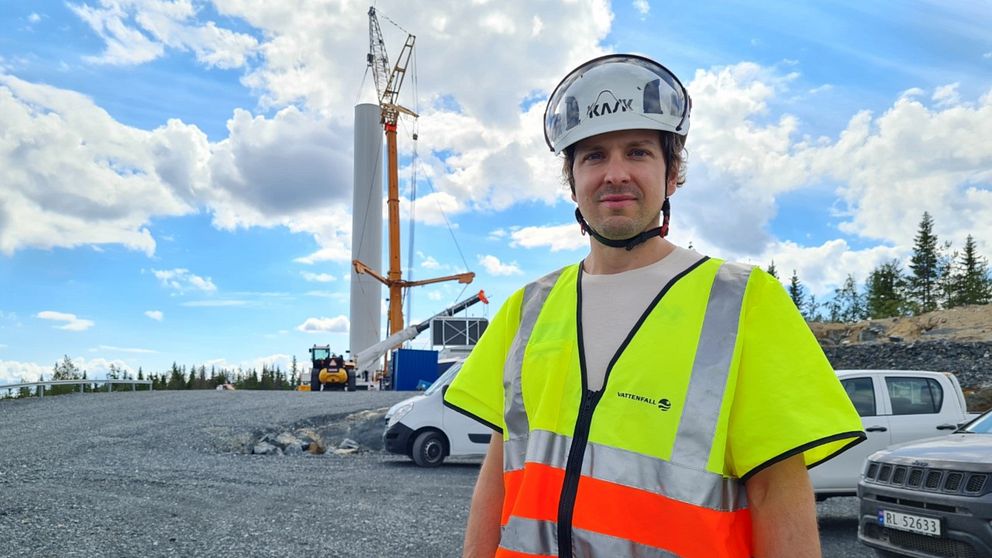 En man med gul och orange reflexväst och vit hjälm står framför byggnationerna av ett vindkraftverk.