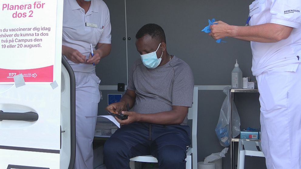 En man sitter på en stol och bär munskydd och grå t-shirt väntar på att få sitt vaccin. En sköterska klädd i vitt till vänster och en sköterska klädd i vitt till höger om mannen.