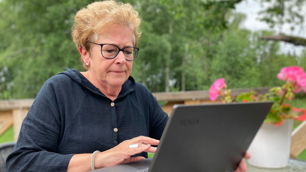 Bild på en kvinna i 60-årsåldern som sitter framför en svart laptop. Hon sitter utomhus på en veranda, i bakgrunden skymtar grönska.