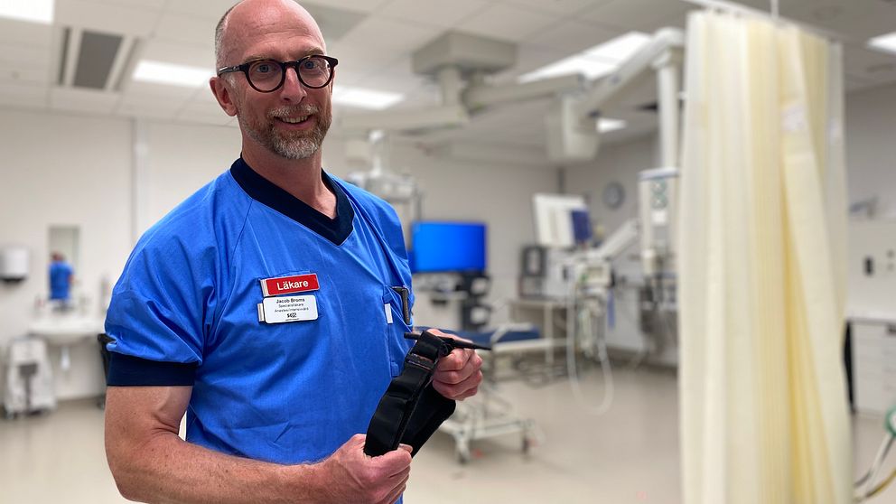 Läkaren Jacob Broms visar upp det avsnörande förband som är bra att använda för att stoppa en blödning. Han har blå kläder, bakgrunden är suddig men han står i ett akutrum på sjukhuset.