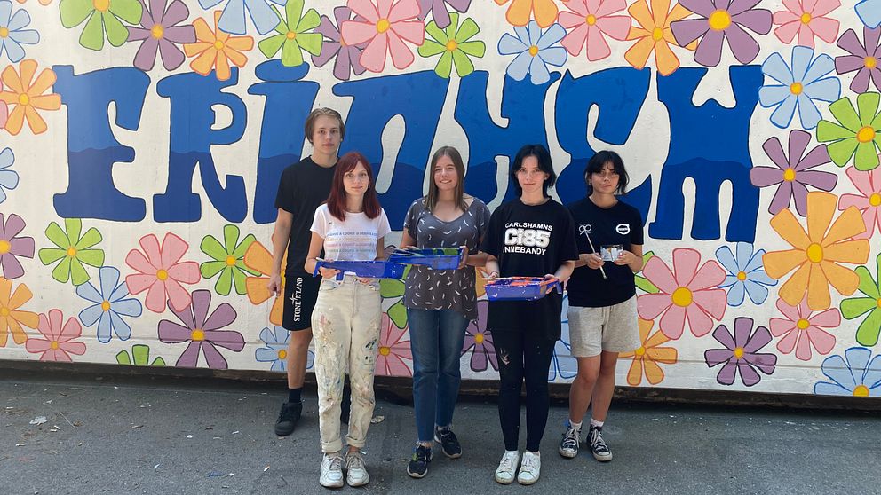 Fem ungdomar framför en färgglad vägg med blommor och stora bokstäver.