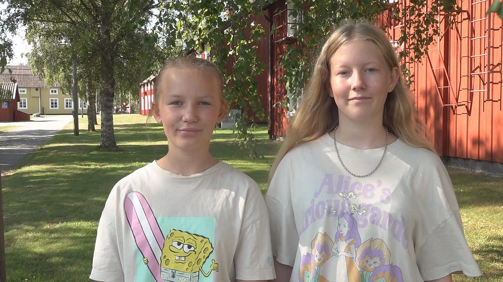 Två unga tjejer med blont hår står bredvid varandra. i Bakgrunden syns röda hus och björkar.