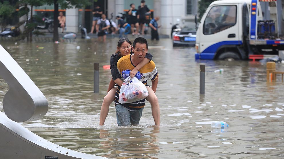 En man bär en kvinna på ryggen i midjehögt vatten på gatan i Kina. Kvinnan är vid medvetande och rynker på näsan. Hon håller i en plastpåse.