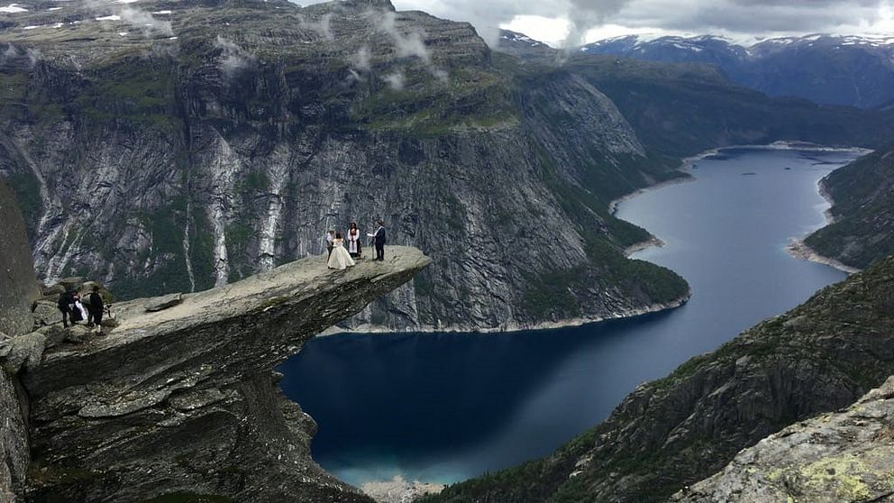 Ett bröllopspar, en vigselförättare och en violinist står längst ut på en klippavsats. Runt dem syns berg och en sjö.