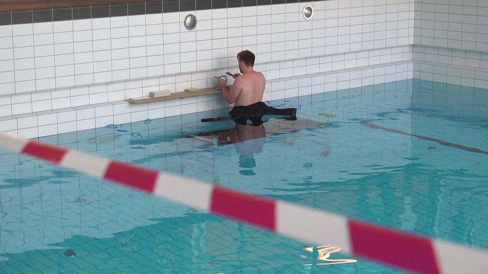 en man i badshorts står vid bortre bassängkanten med ryggen mot kameran och jobbar med kaklet, avspärrningsband i förgrund
