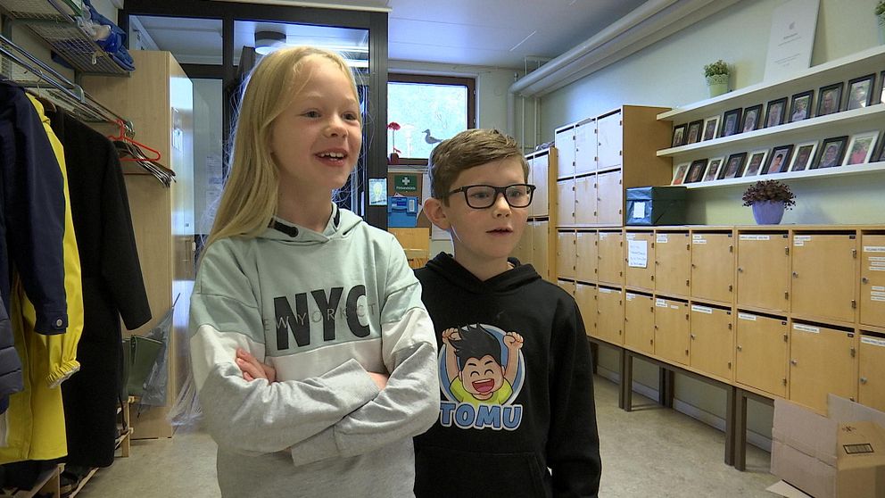 Till vänster står en blond tjejer med armarna i kors och ler, till höger står en brunhårig kille med glasögon. Barnen befinner sig i en skolmiljö med skåp och kläder runt omkring dem.