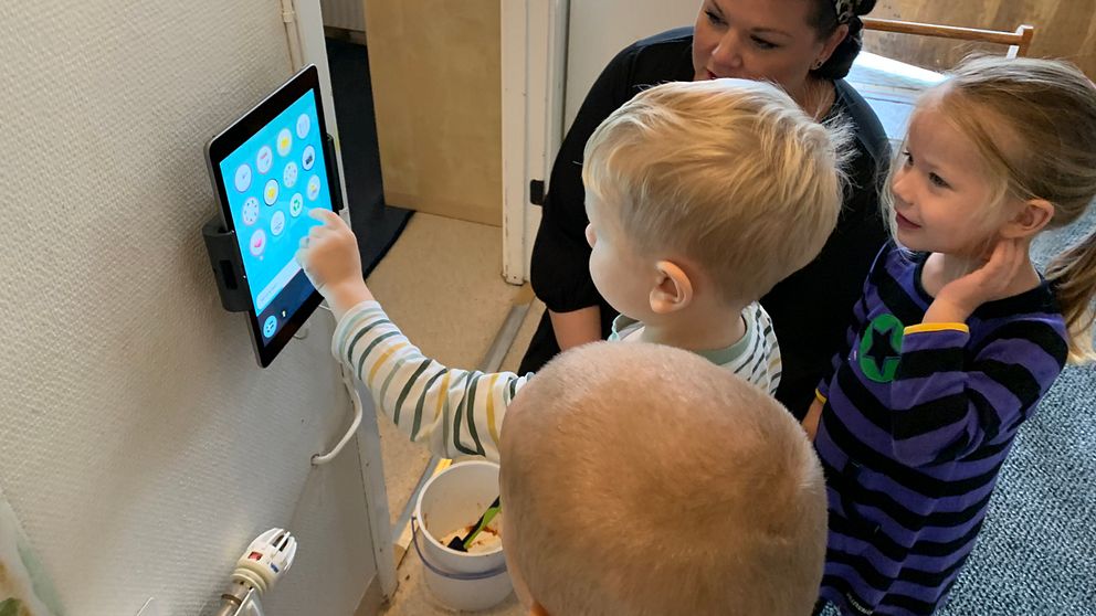 Tre förskolebarn och en vuxen tittar på en surfplatta som sitter på väggen.