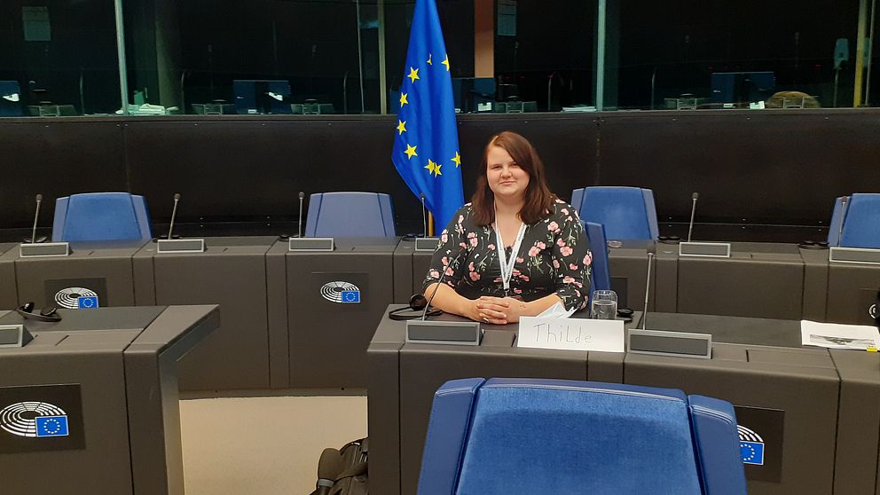 Thilde sitter vid ett bord. bakom sig har hon EU:s flagga.