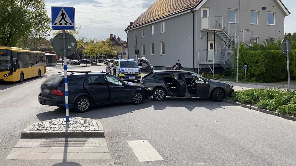 En svart civil polisbil har kört in i en grå bil och tvingat den till kanten strax intill ett övergångsställe. Bildörrarna och bakluckan står öppna. I bakgrunden syns en större polisbil.