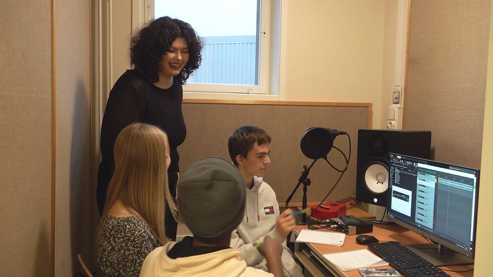 En svartklädd kvinna med lockigt hår står i ett rum där tre unga personer sitter samlade vid en dator. Kvinnan skrattar.