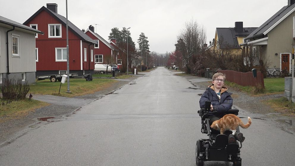 En tonårspojke sitter i en elrullstol, mitt i ett bostadskvarter. På hans ben står en orange katt.