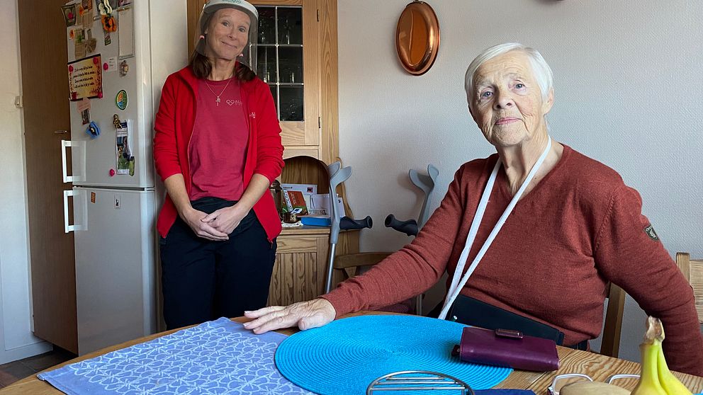 Elisabeth sitter vid sitt köksbord. I bakgrunden står Johanna Karlsson i röd hemtjänstfleece och ansiktsvisir. Elisabeth ser allvarlig ut, Johanna ser glad ut.