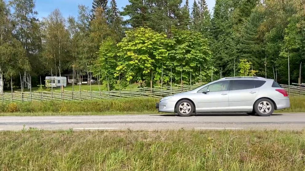 en landsväg i Dalarna, en bil som kör, grön skog i bakgrunden och grönt gräs på vägrenen, i bakgrunden skymtar en husvagn bakom en gärdsgård.
