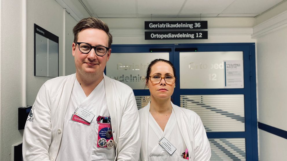 en man till vänster och en kvinna till höger tittar in i kamera, klädda i sjukhuskläder står i en sjukhuskorridor.