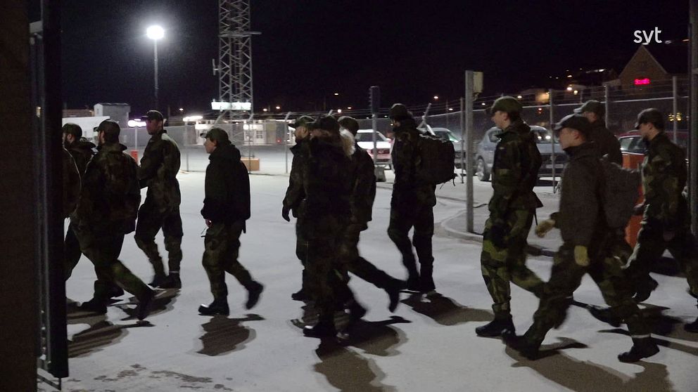 Militärpersonal i vinterkläder går genom en upplyst, snötäckt plats på natten.