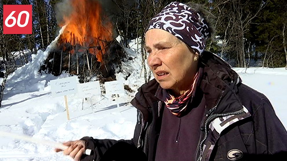 En äldre kvinna står framför en kåta som brinner