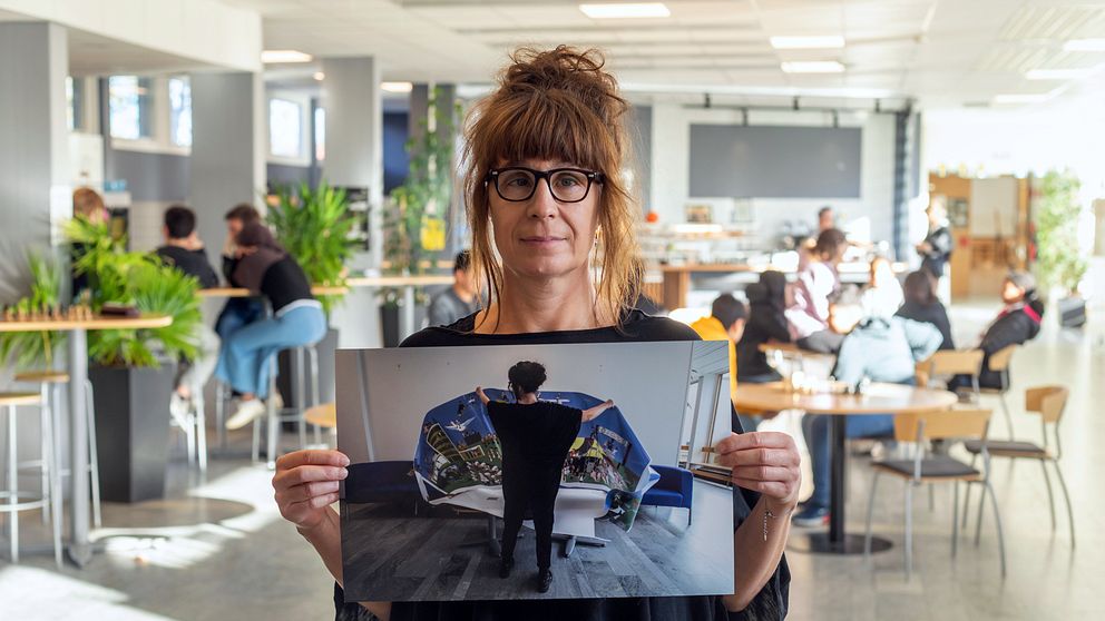 Kvinna i skolmiljö håller upp bild på en kvinna om håller i en affisch