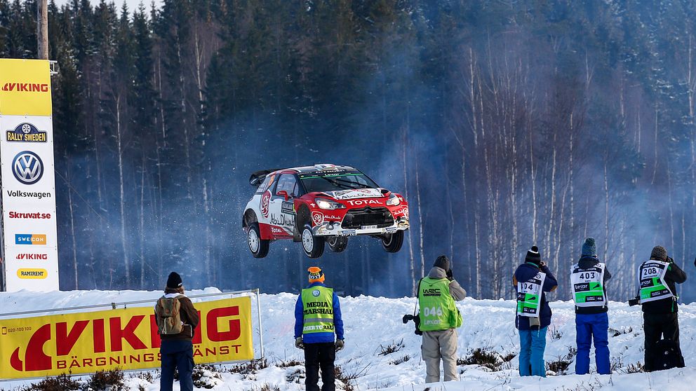 En rallybil i luften efter ett hopp på vinterväg. Vid sidan av vägen funktionärer och pressfotografer