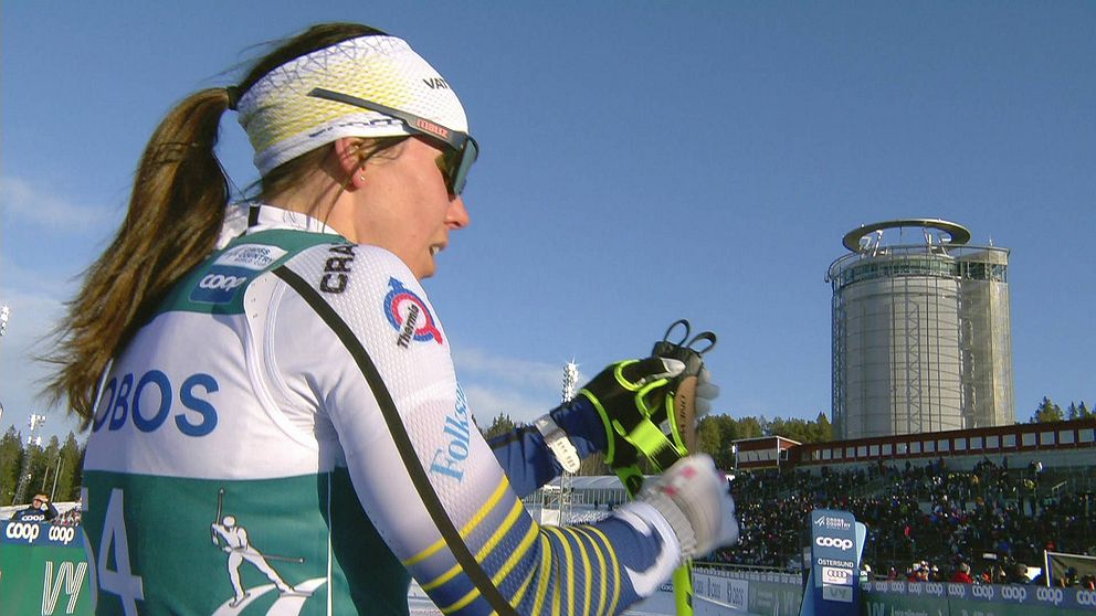 Bild på kvinnlig skidåkare i vit svensk landslagsdress med tornet Arctura i bakgrunden. Från Östersunds skidstadion.