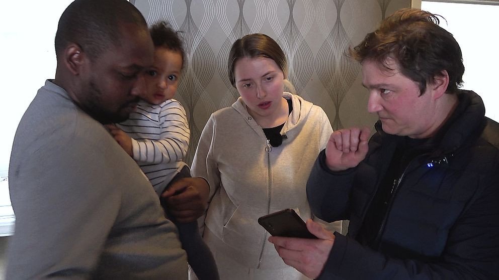 en man med ett litet barn på armen och en kvinna, som pratar med en medelålders man med mobiltelefon