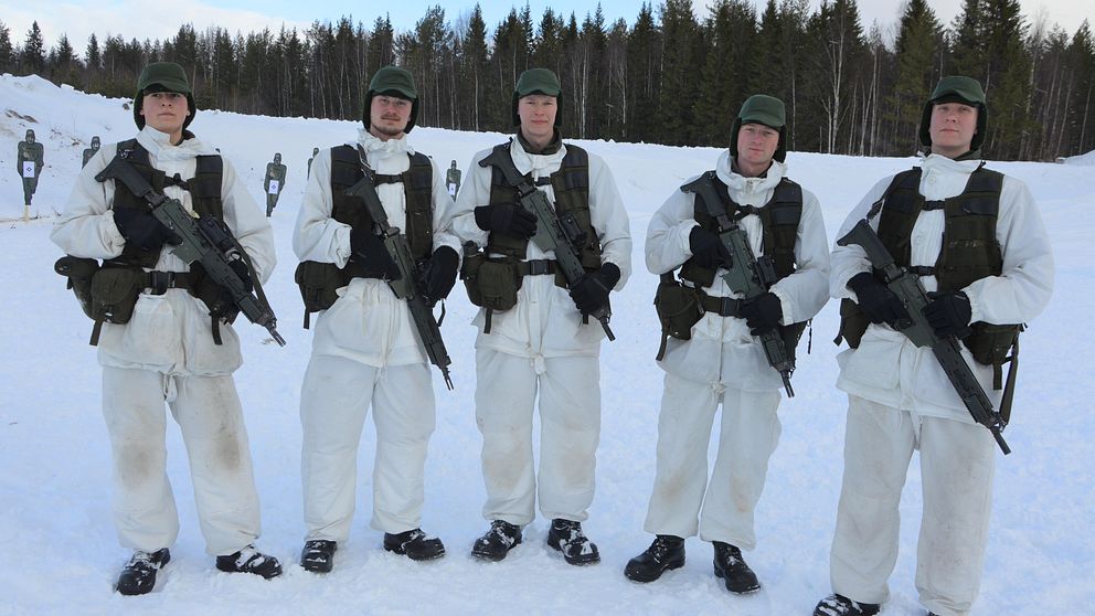 Fem unga militärer i vit vintermuntering står och håller i varsitt automatgevär.