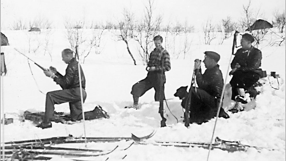 gammalt svartvitt foto på fyra män som sitter och står i snön, skidor bredvid, en håller gevär