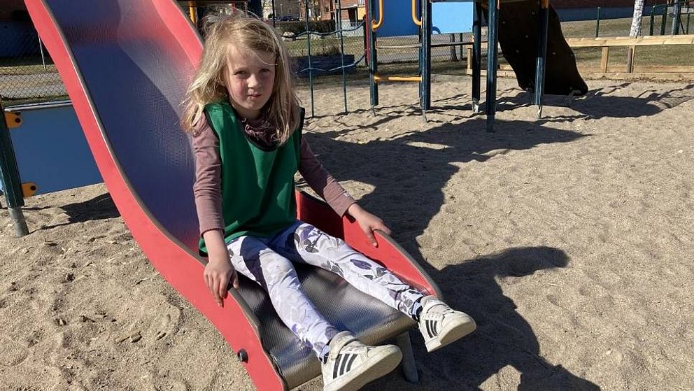 en femårig flicka sitter på botten av en rutschkana i sandtäckt lekpark