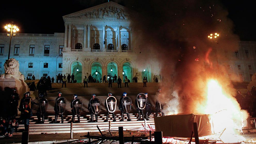 Poliser med kravallutrustning står brevid varandra framför parlamentsbyggnaden. I förgrunden brinner en eld som tänts av demonstranter. foto: scanpix