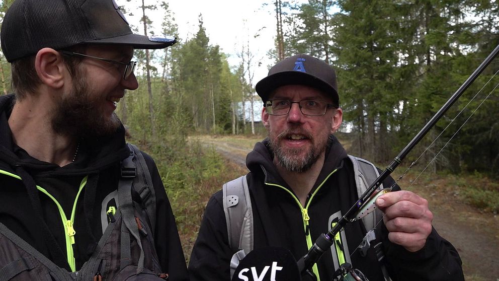 Två män med ett fiskespö står och flinar mot kameran på en skogsväg, sjö skymtar