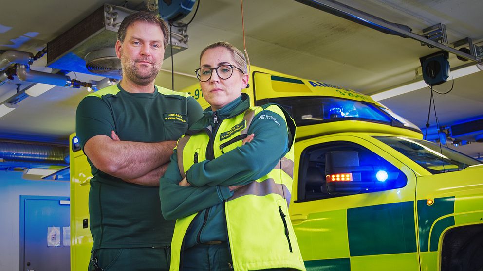 Ambulanssjuksköterskorna Jonas Brindmark och Sofie Smedberg står med armarna i kors framför en ambulans i ambulansgaraget.