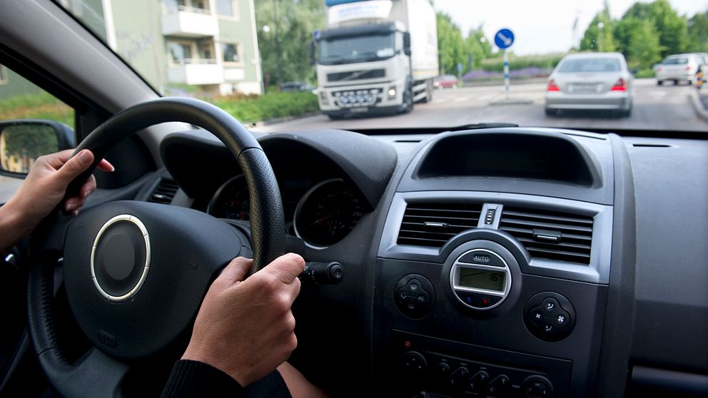 En person kör bil. Man ser händerna på ratten och annan trafik genom vindrutan.