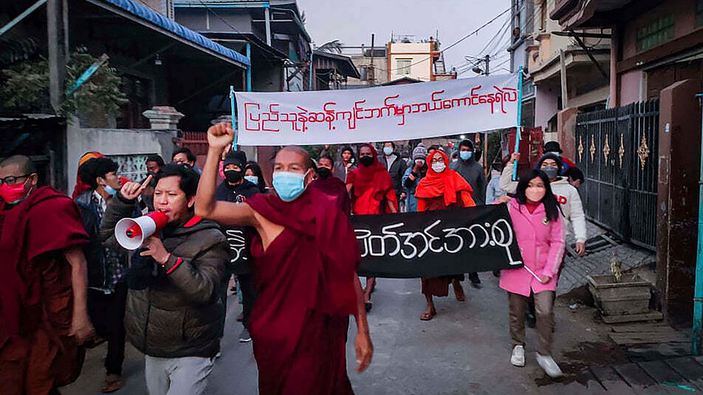 Ungdomsaktivister och buddhistiska munkar deltar i ett protestmöte mot den militära regeringen samtidigt som de håller i en banderoll som lyder på burmesiska: ”Vem vågar stanna på motsatt sida av folkets vilja”, tisdagen den 1 februari 2022, i Mandalay, Myanmar.