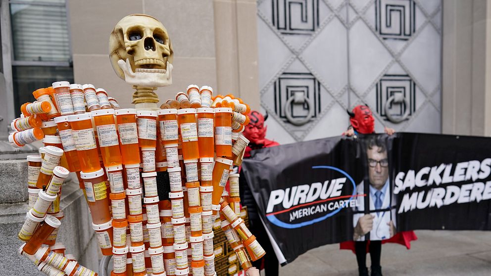 En utställning med en skelettmodell täckt av tomma receptflaskor står i förgrunden, symboliserande opioidkrisen. I bakgrunden protesterar personer i maskerade kostymer med en banner som visar texten ”Purdue American Cartel” och ”Sacklers Murderers”. Bilden fångar en demonstration mot Purdue Pharma och dess koppling till opioidmissbruk.
