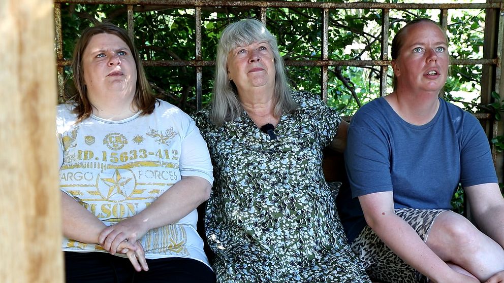 Tre kvinnor sitter på en bänk med en buske bakom.
