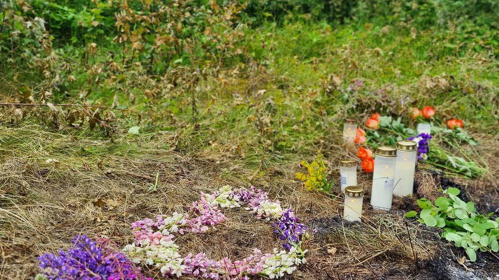 blonkrans och gravljus på marken vid en förkolnad fläck, skog bakom