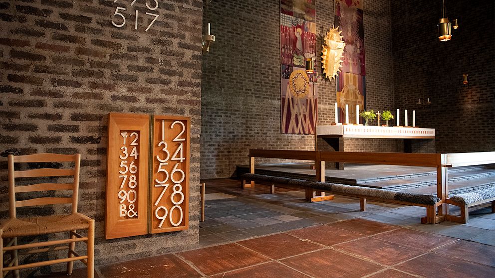 Interiör i en kyrka med altartavlor mot en tegelvägg, en stol, kor och altare med duk och stearinljus.