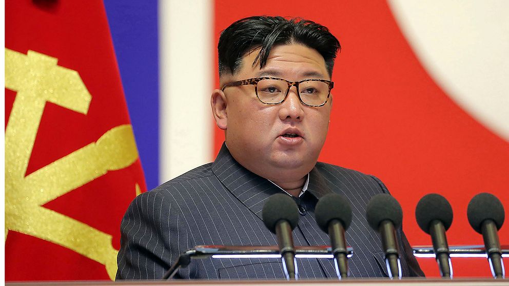 Nordkoreas ledare Kim Jong Un sitter framför en nordkoreansk flagga och flera mikrofoner och håller tal.