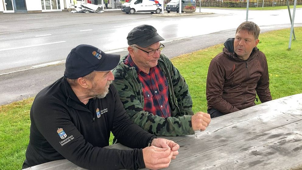 Tre män sitter vid en bord vid en gata. I bakgrunden en mack.