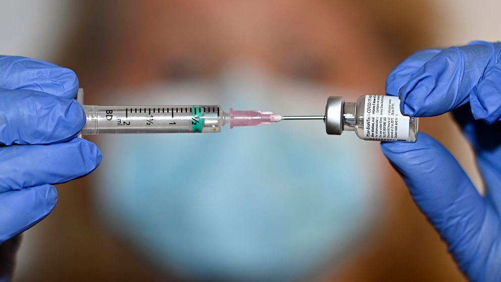 När bild på spruta (nål) i vaccinbehållare, detta är skarp. I bakgrunden ett oskarpt kvinnoansikte, som har ett ljusblått munskydd.