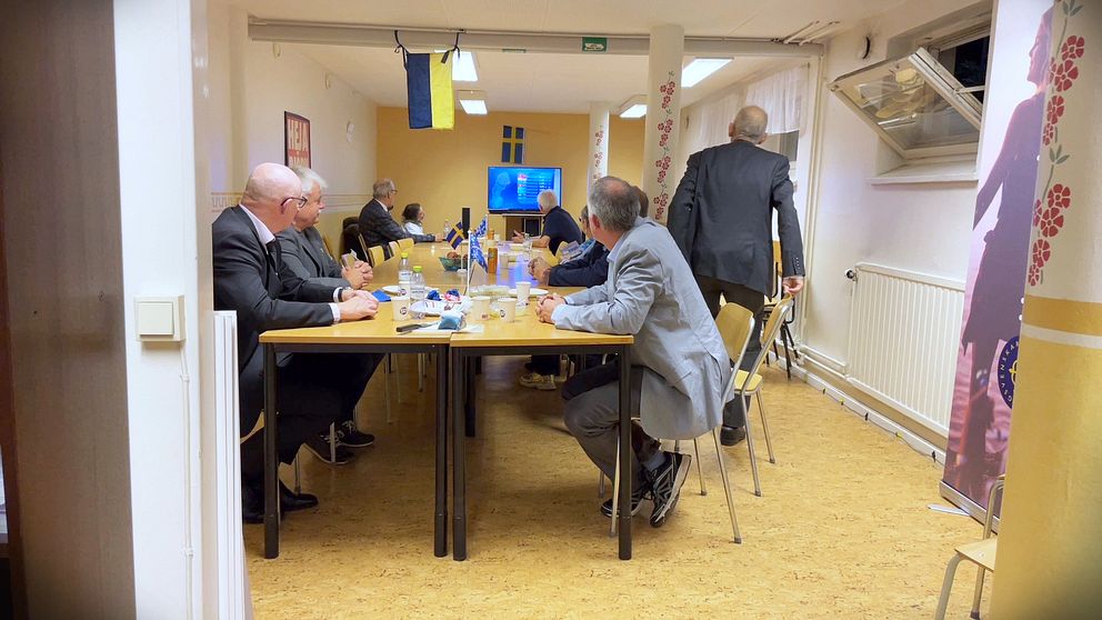 Flera män sitter vid ett avlångt bord för att titta på en tv-apparat som står längst bort i rummet. Tv:n visar SVT:s valvaka.