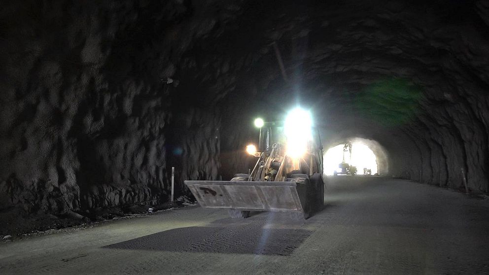 en hjullastare med skopa och lampa på väg i en tunnel, öppningen syns i bakgrunden