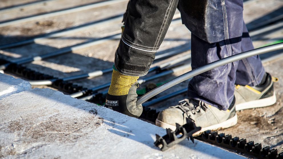 byggarbetsplats, byggnadsarbetare som installerar golvvärme i ett betonggolv, grova kängor, jobbarkläder och handskar. man ser bara fötter, ben och en hand, arbetar framåtböjd nere på golvet.