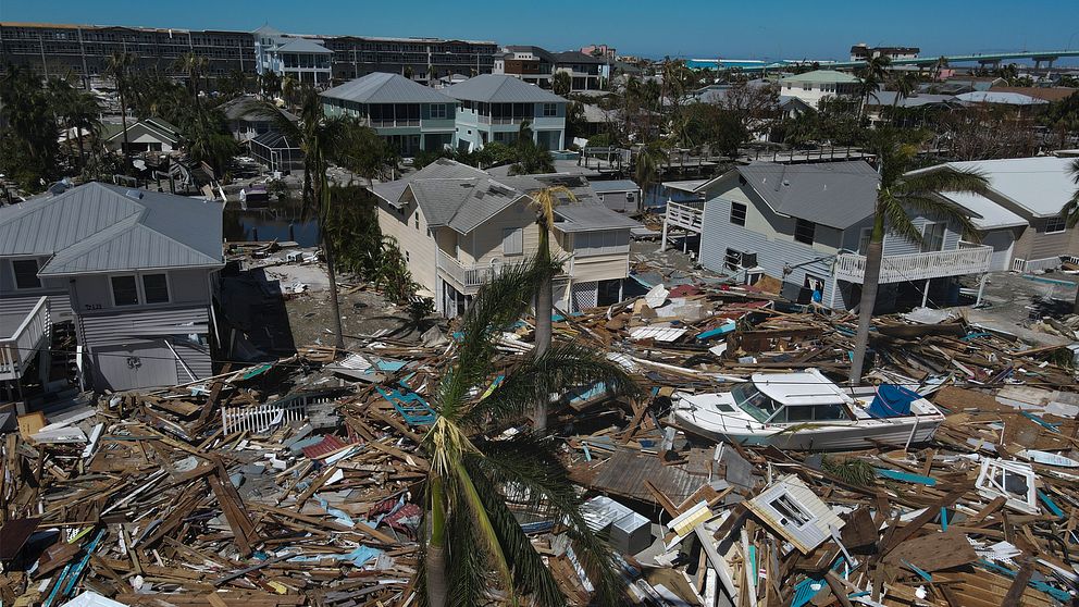 Drönarbild över förstörda byggnader som har dragits med från stranden bredvid hus, palmer och en motorbåt.