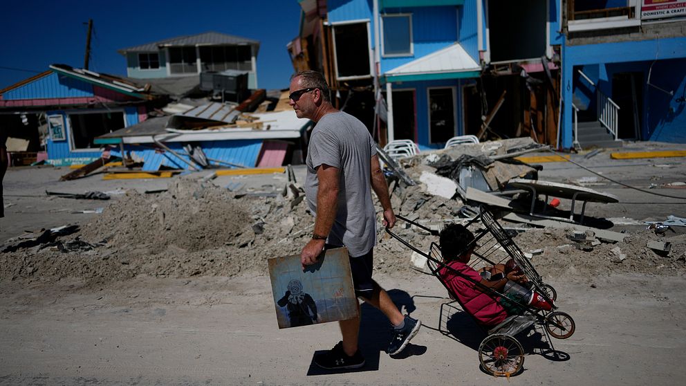 En man går på gatan i Fort Myers Beach. Han bär en målning föreställande en astronaut i ena handen medan han drar en vagn med en pojke i andra handen. Bakom honom syns förstörda hus och byggnader.