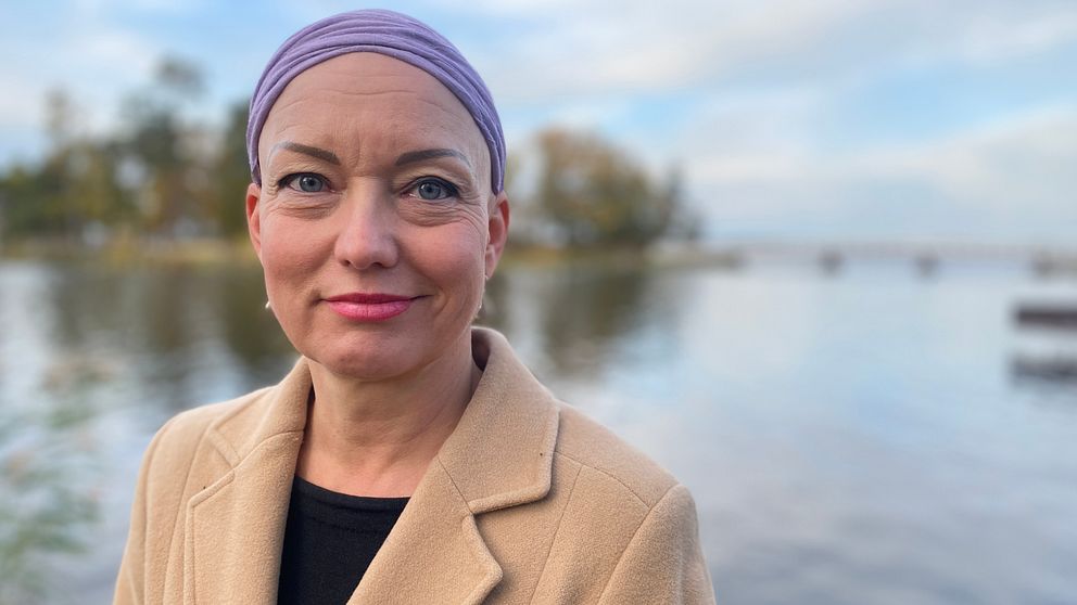 Bild på Carola Forssell, deltidsboende i Mälarbaden utanför Eskilstuna. Hon tittar in i kameran. På sig har en beige kappa med en svart tröja under. På huvudet har hon en lila sjal. Hon har rosa läppstift.